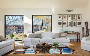 A modern minimalist living room with white furniture, wooden floors, and large windows showcasing a wooden barn outside.