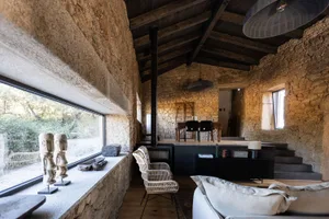 An industrial-style living room with stone walls, a rustic wooden ceiling, and a large window overlooking the outdoors.
