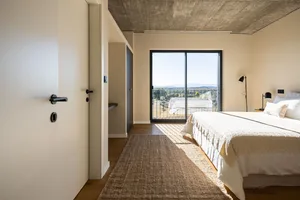 Modern minimalist bedroom featuring concrete walls and wooden flooring, with a view of the surrounding landscape.