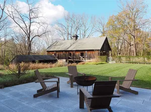 Outdoor deck with wooden chairs and a fire pit surrounded by greenery and a wooden barn in the background.