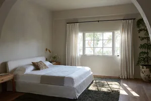A minimalist bedroom featuring a white fabric bed with a plain white headboard, white bedding, and a natural light streaming
