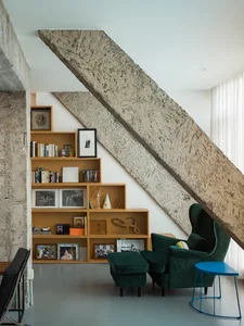 A modern industrial living room featuring a wooden bookshelf and concrete staircase, with a green velvet armchair and ottoman