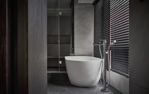 A minimalist modern bathroom with a freestanding white bathtub, concrete floors, and black Venetian blinds on the window, cre