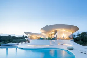 A modern house featuring large glass walls and a concrete structure, with a pool and outdoor seating area.