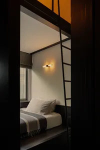 A minimalist bedroom with a single bed, pillows, and a blanket, featuring a wooden beam ceiling and a ladder.