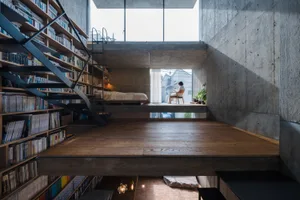 Minimalist residential interior featuring concrete and wood materials, with a person sitting at a window.