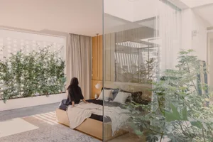 A modern bedroom featuring a wooden platform bed with white bedding and a hanging light fixture, surrounded by green plants.