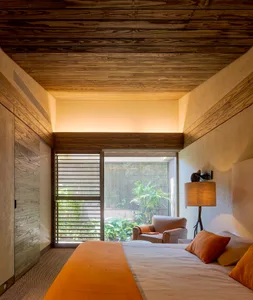 A modern bedroom with a wooden ceiling, orange bedding, and a view of a tropical garden.