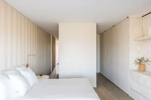 A minimalist bedroom with white walls, a bed with white bedding, and a wooden headboard.