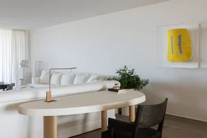 A minimalist living room with a white sofa, desk, and chair, accented with a yellow and blue painting.