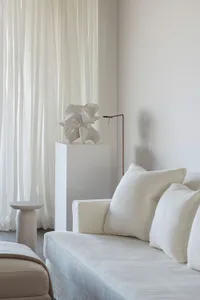 A minimalist living room featuring a white fabric couch, a lamp, and a decorative sculpture with soft lighting and white curt