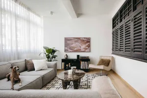 A modern minimalist living room with a grey sectional sofa, a marble coffee table, a beige armchair, and a large shuttered wi