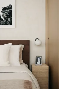 A minimalist bedroom featuring a wooden headboard and a concrete nightstand with a small framed photo and a white lamp.