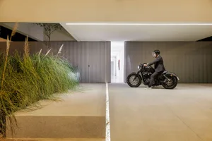 一位身穿全黑服裝的男子騎著黑色摩托車，在現代室內地板上行駛，旁邊有一排綠色樹籬。