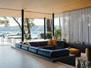 A minimalist living room with a blue sectional sofa, wooden flooring, and large windows overlooking a waterfront view.