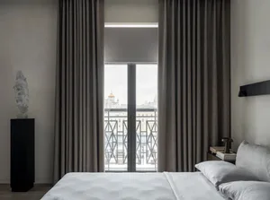 A modern, minimalist bedroom featuring a grey bed and curtains with a cityscape view through the window.