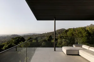 Modern outdoor seating area with glass walls and concrete floor, overlooking greenery and hills.