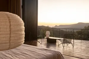 A modern bedroom featuring a wooden floor with a large window overlooking mountains at sunset.