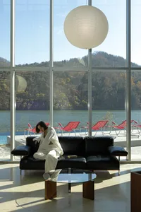 A modern interior featuring a glass window frame, with a black sofa, red lounge chairs, and a large white light fixture.