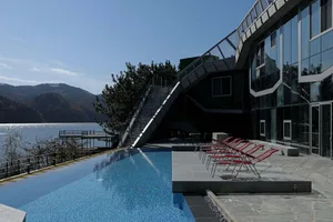 Modern house with glass and metal exterior overlooking a lake, featuring an outdoor pool with red lounge chairs.