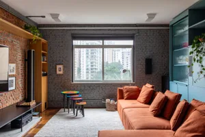 A spacious, industrial-style living room with brick and concrete walls, an orange sectional sofa, a large window, and a mix o