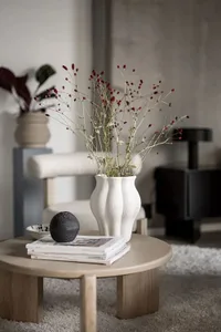 A minimalist decor table scene featuring a ceramic vase with red berries and greenery, placed on a round wooden table with a 