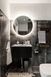 A modern minimalist bathroom with concrete walls and floor tiles, a floating vanity, and a round illuminated mirror.