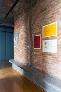 An exhibition space with exposed brick walls and a concrete bench, featuring colorful framed artworks on the wall.