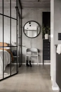 A minimalist bedroom with concrete walls, a glass partition, and a round mirror.