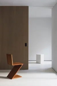 A minimalist space featuring a wooden zigzag chair and a white cube on a polished concrete floor.