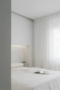 A minimalist bedroom with white walls, curtains, and bedding, featuring a lighted alcove above the bed.