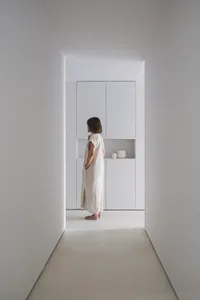 A minimalist white interior with a single person standing in a narrow hallway.