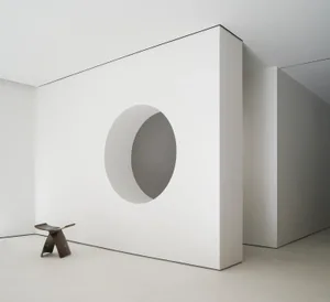 A minimalist white concrete room with a circular cutout in the wall and a wooden stool in a white interior setting.
