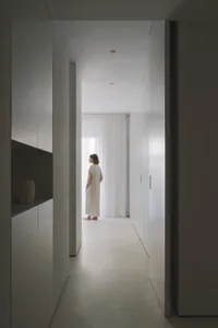 A minimalist hallway with white walls and flooring, featuring a person standing by a window.