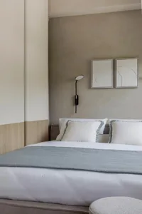 A minimalist bedroom with gray and white bedding, framed abstract art, and a modern fabric ottoman.