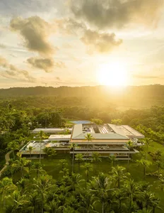 A modern villa with glass walls, concrete structures, and wooden accents surrounded by lush greenery during sunset.
