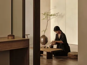 A person sitting cross-legged on the floor performing tea ceremony in a serene, minimalist room with wooden furniture and cer
