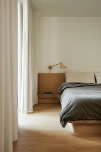 A minimalist bedroom featuring a wooden floor, bed with a wooden headboard, and simple wall-mounted lamp.