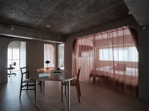 A minimalist residential interior featuring concrete walls and floors, with a table, chairs, and a bed.