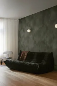 A modern minimalist living room with a green velvet sofa, a white table lamp, and a green textured wall.