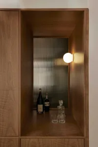 A modern bar shelf made of wood with a spherical wall-mounted light fixture, bottles of wine and liquor, and glasses.