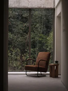 An industrial-style living room with a brown fabric armchair, metal frame, and glass window overlooking a forest.