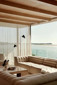 A minimalist living room with a couch, coffee table, floor lamp, and sheer curtains overlooking a body of water.