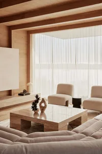 A modern minimalist living room with neutral-toned furniture, clean lines, and a large window with sheer curtains allowing na