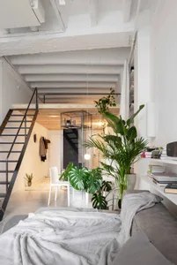 A modern industrial loft space featuring concrete stairs, green plants, a sofa with books, a hanging light fixture, and a spi