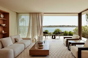 Luxurious neutral-toned living room featuring a comfortable sofa and chairs with a large window overlooking serene waters.