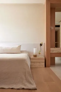 Minimalist bedroom with a wooden floor, beige bedding, and a wooden nightstand with a candle and a vase of flowers.