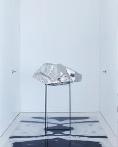 A modern metallic aluminum sculpture displayed on a black stand in a minimalist room.
