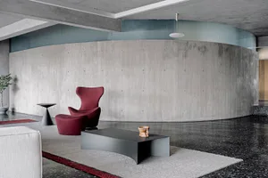A modern living room with concrete walls and flooring features a white sofa and a red chair.