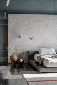 A minimalist bedroom with a concrete wall, a bed, a wall lamp, and decorative vases.